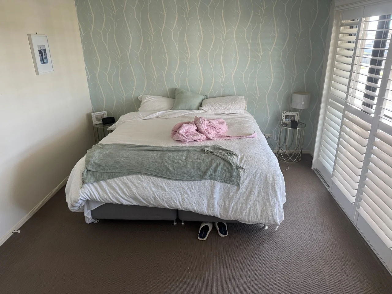 A lived-in bedroom photographed on a phone: an unmade bed, side tables and slippers on the carpet.